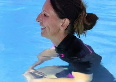 Femme souriante pratiquant la sophrologie aquatique en piscine chauffée, bras détendus en flottaison, vêtue de combinaison