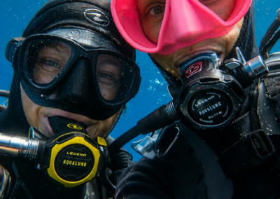 deux femmes en plongée sous marine, avec l'une un masque rose, l'autre un masque noire, elles sont joyeuses