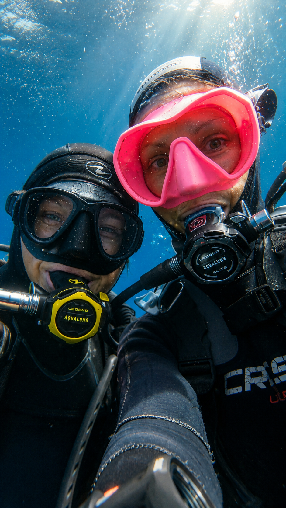deux femmes en plongée sous marine, avec l'une un masque rose, l'autre un masque noire, elles sont joyeuses