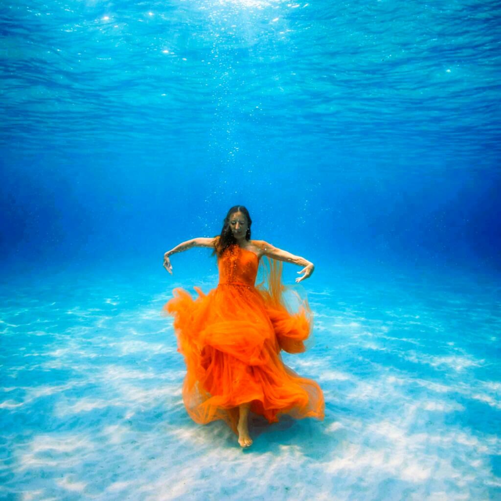 Femme en robe orange flottante sous l'eau en sophrologie aquatique, bras écartés en posture de détente et lâcher-prise en